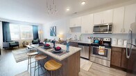 This thoughtfully designed single-floor residence offers 1 bedroom and 2 bathrooms within a compact yet efficiently laid-out floor plan. The open-concept kitchen and living area serves as the heart of the home, where a granite-topped island with dark wood cabinetry and bar seating anchors the space. The kitchen is fully equipped with stainless steel appliances, including a refrigerator, range, microwave, and dishwasher, complemented by crisp white upper cabinets and a classic subway tile backsplash. A modern pendant chandelier adds an elegant touch overhead. Light wood-style flooring flows throughout the main living area, which features large windows that invite natural light and offer outdoor views. The living room provides a comfortable, open setting with neutral walls and ample space for relaxed seating arrangements. The bedroom is carpeted for comfort, bathed in natural light through a large window, and includes a ceiling fan. A dedicated walk-in closet with built-in shelving offers generous storage. The two bathrooms are finished with wood-toned vanities and granite countertops, one featuring a tub-shower combination. The overall ambiance is modern, clean, and functional.