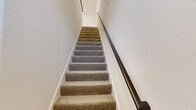 This thoughtfully designed two-floor residence offers a clean, modern aesthetic with light wood-tone flooring and crisp white walls throughout. The main living level is bright and open, anchored by a spacious living area with recessed lighting, a ceiling fan, and large sliding glass doors that lead to a private balcony with outdoor views — filling the space with abundant natural light. The kitchen is a standout feature, outfitted with white shaker cabinetry, quartz countertops, a large peninsula with an undermount sink, and a full suite of stainless steel appliances including a refrigerator, range, microwave, and dishwasher. A dedicated laundry area is conveniently tucked within the kitchen space. The upper floor hosts the primary bedroom, which offers a generous footprint, a ceiling fan, a bright window with outdoor views, and direct access to a well-sized walk-in closet with built-in shelving. The full bathroom is finished with light wood-tone flooring, a white vanity with a gold-framed mirror, and a window for natural ventilation. Carpeted stairs with a dark wood handrail connect the two levels seamlessly. The overall ambiance is clean, airy, and move-in ready.