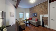 This single-floor space masterfully combines historic architectural character with modern comfort. The living room is the standout centerpiece, featuring a dramatic exposed brick accent wall, a bold dark wood ceiling beam, and rich hardwood flooring throughout. Multiple windows invite generous natural light, while a comfortable seating arrangement — including a full sofa and lounge chairs — anchors the space around a dark wood coffee table, creating a warm and inviting atmosphere.

The kitchen is well-appointed with warm wood cabinetry extending to the upper walls, dark stone countertops, and a full suite of stainless steel appliances including a range, refrigerator, and dishwasher. A wood beam detail continues here, reinforcing the cohesive character of the space.

The unit includes multiple bedrooms, each finished with the same rich hardwood floors and crisp white walls. One bedroom benefits from multiple windows on two walls, flooding the room with natural light. Another features mirrored sliding closet doors and a sleek, contemporary design.

The bathroom is clean and efficiently designed, with white tile, a pedestal sink with chrome fixtures, and a framed mirror, offering a bright, functional space. Throughout, the interplay of original architectural details and updated finishes gives this residence a distinctive, urban character.