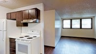 This single-floor space offers a straightforward and functional layout, connecting a kitchen area to an adjoining living space in an open, practical arrangement. The kitchen is equipped with a white electric range and overhead vent hood, complemented by dark-stained wood cabinetry that provides ample storage above. The rich, dark hardwood flooring runs continuously throughout, lending a sense of warmth and cohesion between the kitchen and the connected room. A textured ceiling adds a classic touch to the interior. The adjoining room benefits from a double-pane window that invites natural light, creating a bright and welcoming atmosphere. The neutral wall tones throughout keep the space feeling clean and versatile, offering a blank canvas ready for personalization. The overall layout is compact yet efficient, making practical use of every area. The seamless flow between the kitchen and the neighboring room makes the space well-suited for everyday living, with natural light and consistent flooring tying the areas together into a unified whole.