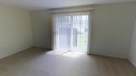 This two-floor residential space offers a practical and comfortable layout designed to maximize light and livability. The living area is a standout feature, highlighted by a large sliding glass door dressed with vertical blinds that floods the room with natural light and provides direct views of the lush green outdoor surroundings. The neutral-toned carpeting throughout creates a clean, cohesive look that complements the simple, understated wall finishes. The open feel of the main living space lends itself well to a variety of furniture arrangements and uses. The overall design is straightforward and functional, with a light color palette that enhances the sense of openness throughout. Spanning two floors, the layout offers separation between living and private spaces, a practical feature for everyday living. The combination of ample natural light, outdoor views, and a clean, neutral interior creates a welcoming and versatile environment suited to a range of lifestyles.