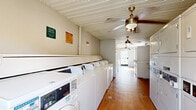 Located at 840 S Grand Hwy in Clermont, Florida, this single-floor community laundry facility offers a practical and well-organized space designed to accommodate high-volume laundry needs. The layout is elongated and efficiently arranged, with a row of commercial-grade top-loading washing machines and dryers lining one wall, complemented by a mix of front-loading and stacked washer-dryer units to maximize capacity. Warm hardwood flooring runs the length of the space, providing a clean and durable surface underfoot. Multiple ceiling fans are installed throughout, ensuring consistent airflow and comfort during use. A built-in granite-topped folding counter runs along one section of the wall, offering a convenient surface for sorting and folding laundry. Natural light filters in through windows and a glass-panel entry door at the far end, contributing to a bright and welcoming atmosphere. The walls are finished in neutral tones, and the beadboard-style ceiling adds a subtle architectural detail. Posted signage including laundry room rules and safety cautions reflects an attentive, organized facility management approach. The space is straightforward and functional, purpose-built to serve the laundry needs of a residential community.