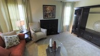 This single-story living space is designed with comfort and elegance in mind. The living room serves as the focal point, centered around a classic fireplace with ornate black metalwork screening, which adds a traditional yet refined character to the room. Above the fireplace, a framed artwork provides a striking visual accent against the soft, neutral-toned walls.

The room benefits from abundant natural light, with multiple windows dressed in floor-length draping curtains that lend a sense of height and sophistication. Plush wall-to-wall carpeting in a warm beige tone contributes to the cozy ambiance underfoot.

Furnishings include a cushioned accent chair with a coordinating throw pillow, positioned alongside a dark wood side table. A circular glass-top coffee table rests atop a textured area rug, adding a contemporary contrast to the traditional elements of the space. A dark wood media cabinet occupies one corner, offering both display and storage functionality.

The overall aesthetic blends traditional and transitional design elements, creating a balanced, livable space that feels both welcoming and well-appointed.