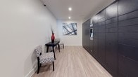 This well-appointed package room is designed with both functionality and aesthetics in mind. The space is anchored by a large bank of dark, smart parcel storage lockers equipped with a digital interface panel, offering secure and convenient package retrieval. The lockers span nearly the full length of one wall, providing ample storage capacity for residents or building occupants.

The flooring throughout is a light wood-style plank that lends warmth and a contemporary feel to the room. Crisp white walls and recessed ceiling lighting create a bright, clean atmosphere. A small seating area features a dark accent table and two patterned upholstered chairs, adding a welcoming touch to what is primarily a utilitarian space. An abstract wall art piece in cool gray tones completes the decor, elevating the room's visual appeal.

The layout is open and easy to navigate, with a clear corridor leading toward a door with a full-length glass panel that connects to an adjacent stairwell or hallway. Additional doors provide further access points within the building. Ventilation grilles are integrated discreetly into the walls and floors, ensuring comfortable air circulation. Overall, this space combines practical infrastructure with thoughtful design details.