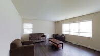 This single-floor space offers a clean, functional layout designed with simplicity and practicality in mind. The living area serves as the focal point of the space, featuring rich dark hardwood flooring that contrasts crisply against the neutral, light-toned walls and bright white ceiling. Multiple windows fitted with horizontal blinds allow natural light to filter generously into the room, creating a warm and inviting atmosphere throughout the day. The open layout of the living area provides a comfortable sense of spaciousness, with ample room for seating and everyday living. The neutral color palette throughout lends itself well to a variety of décor styles, making the space highly adaptable. Baseboard heating runs along the walls, providing an efficient and unobtrusive climate solution. The overall ambiance of the space is understated and versatile — ideal for those seeking a straightforward, move-in-ready environment with solid foundational features including durable flooring and good natural light. The single-floor design ensures ease of accessibility and a cohesive flow throughout the entire living space.