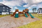 Located at 100 Oakbrook Drive in McDonough, Georgia, this well-appointed outdoor community amenity space offers a thoughtfully designed environment for residents of all ages. The centerpiece of the grounds is a substantial wooden playground structure featuring multiple green slides, elevated platforms, a suspension bridge, and climbing elements, all set over a cushioned gravel surface for safety. The playground is accessible via intersecting concrete walkways that wind throughout the property, connecting key areas and encouraging outdoor activity.

The surrounding landscape is carefully maintained, with manicured lawns, ornamental shrubs, and mulched garden beds that add visual interest and curb appeal. Tall Georgia pines frame the backdrop, providing a natural buffer and a sense of seclusion. A community pool area with an enclosed patio, shaded umbrellas, and a brick clubhouse building is visible along one pathway, offering a resort-style retreat for residents.

The residential buildings themselves display a modern architectural style with a bold navy and white exterior, featuring balconies, large windows, and contemporary paneling. Seating benches are positioned along the walkways, inviting residents to enjoy the open-air surroundings. Ample parking and well-lit pathways contribute to the accessibility and functionality of this vibrant community outdoor space.