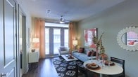 This single-floor space showcases an open-plan living and dining area designed for both comfort and functionality. The living area is anchored by rich, dark hardwood flooring and features a ceiling fan that adds both practicality and a subtle architectural focal point. Large glass French doors flood the space with abundant natural light and provide direct access to an outdoor balcony, framed by warm, sheer orange curtains that add a soft, inviting glow to the room.

The living area is well-proportioned, accommodating a plush sofa and accent chair arranged around a rectangular coffee table, all set atop a bold patterned area rug that grounds the seating arrangement. A vibrant red abstract canvas artwork on the wall serves as a striking decorative focal point, complemented by decorative branches and accent lamps that enhance the warm, contemporary ambiance.

Adjacent to the living space, the dining area features a round white table suited for intimate gatherings. The open layout between these two zones creates a seamless flow, making the overall space feel connected and spacious. The neutral wall tones throughout contribute to a clean, modern aesthetic.