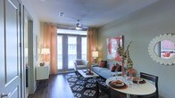 This single-floor space showcases an open-plan living and dining area designed for both comfort and functionality. The living area is anchored by rich, dark hardwood flooring and features a ceiling fan that adds both practicality and a subtle architectural focal point. Large glass French doors flood the space with abundant natural light and provide direct access to an outdoor balcony, framed by warm, sheer orange curtains that add a soft, inviting glow to the room.

The living area is well-proportioned, accommodating a plush sofa and accent chair arranged around a rectangular coffee table, all set atop a bold patterned area rug that grounds the seating arrangement. A vibrant red abstract canvas artwork on the wall serves as a striking decorative focal point, complemented by decorative branches and accent lamps that enhance the warm, contemporary ambiance.

Adjacent to the living space, the dining area features a round white table suited for intimate gatherings. The open layout between these two zones creates a seamless flow, making the overall space feel connected and spacious. The neutral wall tones throughout contribute to a clean, modern aesthetic.