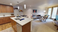 This single-floor space showcases an open-concept layout that integrates the kitchen, dining, and living areas into one cohesive and functional environment. The kitchen features a central island with a white countertop, an undermount stainless steel sink, and warm wood cabinetry. A stainless steel dishwasher is integrated into the island, and pendant lighting hangs above, providing focused task illumination. A wall-mounted shelf adds display and storage opportunities near the kitchen.

The dining area sits adjacent to the kitchen, accommodating a small table and chairs, making it ideal for everyday meals. Flowing seamlessly beyond, the carpeted living area is generously sized, furnished with a sectional sofa accented with colorful throw pillows and complemented by a low-profile coffee table. A piece of vibrant wall art adds a pop of color against the neutral gray walls.

Throughout the space, recessed lighting and pendant fixtures ensure well-distributed illumination, while the neutral tones of the walls and carpet create a calm, cohesive aesthetic. The open floor plan promotes an airy, spacious feel throughout.