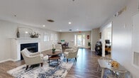This single-floor community space is designed to provide a comfortable and functional gathering environment. The main lounge area is anchored by a classic white-mantled fireplace, creating a warm focal point for the room. Rich hardwood-style flooring flows throughout, complementing the neutral, light-toned walls and recessed lighting that illuminate the space evenly. The seating arrangement features a curated mix of tufted accent chairs and coordinating furniture grouped around a round wooden coffee table, all set atop a blue-toned area rug that adds a touch of color and definition to the space. A wall-mounted flat-screen television and a wooden media console provide additional entertainment amenities. Decorative accents, including potted plants, floral arrangements, and tasteful wall art, contribute to the inviting ambiance. A dining area with a small table and chairs is positioned toward the rear of the space, near a set of French doors that introduce natural light and views of the exterior. Adjacent to the main lounge is a dedicated workstation area furnished with a round marble-top table, seating, and a desktop computer, making it suitable for business or administrative use. The space also includes additional utility areas and accessible entry points, making it a well-rounded and versatile community hub.