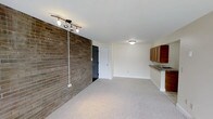 This single-floor space offers a straightforward and functional layout with a blend of classic and practical design elements. The main living area is defined by a striking full-height exposed brick accent wall that adds texture and character to the otherwise clean, neutral interior. Track lighting mounted on the ceiling provides directional illumination, complementing the standard flush-mount fixture positioned toward the center of the space.

The flooring throughout the main area consists of neutral-toned carpet, contributing to a quiet and comfortable underfoot feel. White walls and white trim keep the space feeling bright and open, with a standard white panel door providing access to an adjoining room or closet.

Along one side of the space, a compact kitchen area is partially visible, featuring warm wood-toned cabinetry, a countertop with a built-in sink, and a peninsula-style divider that subtly separates the kitchen from the living area. The white ceiling is smooth and uniform throughout, maintaining a clean and cohesive aesthetic. Overall, the space presents a modest yet practical environment suited for everyday living.