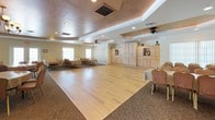 This single-story event venue is designed to accommodate a variety of gatherings, offering multiple interconnected spaces that blend functionality with classic elegance. The main ballroom is a standout feature, boasting a large hardwood dance floor, tiered tray ceilings with warm copper-toned accents, recessed lighting, and ceiling fans. Built-in cabinetry with display shelving lines one wall, adding a refined touch to the space.

Adjacent dining areas feature patterned carpeting, round and rectangular banquet tables with linen coverings, and comfortable upholstered chairs. Tray ceilings with decorative finishes, chandeliers, and ceiling fans carry throughout these rooms, creating a cohesive aesthetic. Large windows and glass doors allow natural light to filter in, enhancing the venue's welcoming ambiance. A wall-mounted projection screen is present for presentations or multimedia use.

A separate room with its own tray ceiling and exterior door access offers a more intimate setting for smaller events or breakout use. The fully equipped catering kitchen features light wood cabinetry, dark granite countertops, tile flooring, and white appliances including a refrigerator, dishwasher, microwave, and oven, providing comprehensive food service support for events.