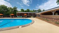 Located at 200 Barnes St in Carrboro, NC, this outdoor community amenity space offers a well-appointed environment designed for relaxation and recreation. At its centerpiece is a generously sized oval swimming pool with clear blue water, surrounded by an expansive concrete sundeck furnished with teal lounge chairs and vibrant orange shade umbrellas — creating a resort-like atmosphere perfect for warm Carolina days. The pool area is enclosed by a classic wrought-iron fence with brick pillars, providing both security and a polished aesthetic. Adjacent to the pool, a covered wooden pavilion with a ceiling fan offers a shaded gathering space, while manicured brick-edged planting beds add attractive landscaping throughout. Beyond the pool enclosure, the grounds open up to a broad green lawn shaded by mature trees, including towering pines that lend the property a natural, park-like feel. A brick outdoor kitchen or grill station is positioned along a paved walkway, ideal for outdoor entertaining. The surrounding multi-story brick residential buildings are classic in construction and set back among the greenery, contributing to a cohesive community setting. The overall ambiance is inviting, well-maintained, and thoughtfully designed for outdoor living.