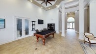 This single-floor community space is designed with a classic, upscale aesthetic that combines elegant architectural details with functional amenities. The grand entryway features soaring ceilings, tall arched windows with transom details, and glass-paneled doors that flood the space with natural light and offer views of an outdoor pool area. Neutral tile flooring in a stone-like finish runs throughout, tying the areas together cohesively.

The main lounge area is anchored by a striking black fireplace with a bold surround and detailed paneled millwork reaching to the ceiling. Deep navy velvet sofas and classic white accent chairs are arranged around a glass-top coffee table, creating an inviting and sophisticated seating area. Abstract artwork adds a contemporary touch against the crisp white walls.

The game room features a full-size billiards table as its centerpiece, complemented by a wall-mounted flat-screen TV, ceiling fan set within a coffered tray ceiling, and recessed lighting. An open corridor lined with tall decorative columns connects the various areas, enhancing the sense of grandeur and flow throughout the space.