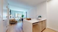Located at 25 Maple Ave in New Rochelle, NY, this thoughtfully designed single-floor residence offers a seamless blend of modern aesthetics and functional living. The open-concept layout connects the kitchen and living area in a fluid, inviting arrangement. The kitchen is anchored by a quartz-topped peninsula with bar seating, light wood cabinetry, a subway tile backsplash, and stainless steel appliances, including a built-in microwave and full-size refrigerator. Light hardwood flooring extends throughout the main living areas, complementing the bright, neutral color palette. The living room benefits from large windows that frame urban views and fill the space with natural light, creating an airy and open atmosphere. A textured accent wall adds visual depth and character to the living space. The bedroom continues the warm, light-filled aesthetic with a generously sized window overlooking the cityscape, providing an abundance of natural light. The bathroom is finished with large-format marble-look tile covering both the walls and floors, paired with a light wood vanity and a soaking tub, offering a clean and refined retreat. Throughout the residence, the consistent use of natural materials and contemporary design elements establishes a cohesive, modern sensibility.