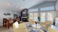 Located at 5909 30th Avenue West in Bradenton, Florida, this single-floor community space offers a thoughtfully designed interior with a warm, coastal aesthetic. The layout includes a formal entrance, multiple hallways connecting distinct zones, and an open main gathering area that benefits from soaring vaulted ceilings that lend a sense of spaciousness well beyond the physical footprint.

The main room serves as the centerpiece of the space, anchored by a striking floor-to-ceiling navy blue accent wall housing a tiled fireplace with a decorative fish school wall sculpture — a nod to the Florida coastal setting. Light wood-look flooring runs throughout, complementing the soft blue-grey walls and creating a cohesive, airy feel. A dining area with a rich wood table sits adjacent, while comfortable cream upholstered seating arrangements invite conversation and relaxation.

Natural light floods the interior through multiple large windows fitted with white blinds and elegant white-framed French doors that open toward an outdoor pool area, seamlessly connecting interior and exterior. A small beverage station and curated coastal wall art add functional and decorative touches throughout. Additional rooms accessible via French door-lined hallways suggest dedicated office or activity spaces, rounding out this versatile and inviting community facility.