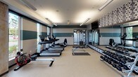 This fitness center offers a comprehensive workout environment designed to accommodate a wide range of exercise routines. The space is defined by a striking interior aesthetic, featuring bold teal and beige horizontal stripes along the walls, complemented by a decorative geometric-patterned accent wall that adds visual depth and a modern flair.

The cardio zone is equipped with multiple treadmills, an elliptical, and a rowing machine, all positioned along the windowed perimeter to allow natural light to fill the space and provide views to the outdoors. A ceiling fan overhead aids in air circulation throughout the room.

On the strength side, the facility includes a multi-function cable machine, an adjustable weight bench, and a full rack of dumbbells spanning a wide range of weights. A dedicated functional training area features stability balls in multiple sizes, medicine balls, a BOSU balance trainer, and open floor space ideal for stretching or mat work. Full-length mirrors along one wall enhance the sense of space while allowing users to monitor their form.

The flooring is a durable, light-toned rubberized surface suited for both cardio and free weight activity. Overhead linear lighting fixtures provide bright, even illumination throughout. A wall-mounted television is positioned for easy viewing from the cardio equipment area, and a water station is conveniently accessible near the entrance.