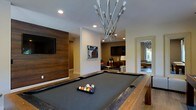 This single-floor entertainment space is designed with a contemporary aesthetic that blends functionality and sophistication. At its centerpiece sits a full-sized billiards table with a charcoal felt surface and a textured dark rail, positioned beneath a striking sculptural chandelier composed of sleek metallic arms radiating outward with exposed bulbs, serving as both a light source and a bold design statement.

The room features a rich wood-paneled accent wall mounting a flat-screen television, adding warmth and texture against the neutral tones of the surrounding walls and ceiling. Recessed lighting complements the overhead fixture, ensuring the space is well-illuminated throughout.

Adjacent to the billiards area, a lounge zone is furnished with comfortable seating, a floor lamp, and an additional wall-mounted television, creating a relaxed setting for socializing. A large decorative mirror on one wall enhances the sense of depth and openness within the space.

A glimpse through a doorway reveals a connected room, suggesting the entertainment area flows into additional spaces. Overall, the design strikes a balance between upscale leisure and modern interior styling.