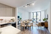 Located at 1 Main St in Miramar, Florida, this thoughtfully designed single-floor residence offers a seamless open-concept layout that efficiently combines the kitchen, dining, and living areas into one cohesive, light-filled space. Light wood-tone flooring runs throughout, complementing the neutral, modern palette.

The kitchen is a standout feature, boasting custom floor-to-ceiling cabinetry in a warm wood grain finish, sleek white quartz countertops, stainless steel appliances including a French door refrigerator and built-in microwave, a dedicated wine cooler, and a central island with bar seating — ideal for both cooking and casual dining. Track lighting above enhances the workspace.

The living area is bright and airy, anchored by expansive floor-to-ceiling glass doors that open to a private balcony with views of lush palm trees and the surrounding community. Sheer white curtains frame the generous natural light that fills the room.

The bathroom features a wide, light wood vanity with ample under-cabinet storage, a white quartz countertop, an illuminated LED mirror, and a full shower-tub combination, all finished in a clean, minimalist style consistent with the rest of the residence. The overall ambiance is modern, serene, and well-curated.