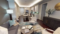 This single-floor space showcases a thoughtfully designed open-concept layout that seamlessly connects the living, dining, and kitchen areas into one cohesive environment. The living area anchors the space with a plush gray sofa, an accent armchair in white, and a round dark coffee table with gold trim, all set atop a vibrant multicolored area rug that introduces teal, gold, and gray tones throughout.

The kitchen is finished with white shaker-style cabinetry, stainless steel appliances, and a teal subway tile backsplash that adds a refined pop of color. A marble-topped island with bar seating serves as both a functional workspace and a natural transition into the dining area, where a pendant light fixture adds an elegant focal point overhead.

A distinctive tray ceiling with integrated LED cove lighting runs across the space, casting a warm, ambient glow that enhances the overall atmosphere. Dark hardwood flooring grounds the design, providing a rich contrast to the light cabinetry and walls. The layout is efficient yet open, making the space feel airy and well-proportioned throughout.