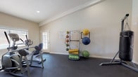 This single-floor fitness center is designed to support a well-rounded workout routine in a clean, functional environment. The space features durable rubber flooring throughout, providing a safe and stable surface for a variety of exercises. Natural light filters in through a glass-paneled door, lending the room an open and welcoming atmosphere while maintaining easy access to the exterior.

The facility is outfitted with cardio equipment, including an elliptical trainer, positioned to make the most of the available floor space. A dedicated equipment rack holds an organized selection of medicine balls in varying sizes and weights, alongside rolled exercise mats, making functional training and stretching readily accessible. Additional free weights and fitness accessories are neatly arranged along the perimeter of the room.

The walls are finished in a neutral off-white tone, contributing to the bright and airy feel of the space. Recessed lighting complements the natural light, ensuring the room remains well-lit throughout the day. Informational notices are posted on the wall near the entrance, providing guidance on equipment use and facility rules. Overall, the fitness center offers a practical and organized setting suited for cardiovascular training, strength work, and flexibility exercises.