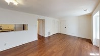This single-story space is characterized by a clean, neutral aesthetic and consistent warm-toned hardwood-style flooring that runs throughout the living areas and bedrooms. The main living area is generously sized, featuring large windows fitted with vertical blinds that allow abundant natural light to fill the room. A ceiling fan with a light fixture adds both function and comfort. An adjacent area, visible through an open layout, connects seamlessly and includes a pass-through window opening, a built-in floor vent for heating, and direct access to the entry door, as well as a closet for storage.

The bedrooms continue the same flooring and neutral wall palette, each equipped with windows dressed in vertical blinds that provide natural light and outdoor views. The rooms offer a versatile, open canvas suitable for varied furniture arrangements.

The bathroom is efficiently designed, featuring square white subway tile lining the tub-shower surround, a soaking tub, a toilet, and a flush-mount ceiling light. The floor is finished with earthy terracotta-toned tiles, complementing the clean white fixtures. A towel bar provides practical storage. Overall, the space presents a functional, light-filled environment with a cohesive and understated design throughout.