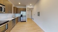This single-floor unit presents a clean, modern aesthetic throughout, characterized by crisp white walls, light wood-style flooring in the main living areas, and an open, functional layout. The entry corridor leads into the heart of the unit, where a smart thermostat and utility panel are conveniently mounted on the wall, reflecting the space's contemporary, tech-forward design.

The kitchen is well-appointed with warm wood-toned cabinetry, light countertops, and a full suite of stainless steel appliances, including a refrigerator with ice maker, electric range, built-in microwave, and dishwasher. Recessed lighting illuminates the space efficiently.

The living area is bright and airy, benefiting from large windows that frame views of surrounding greenery and flood the room with natural light. A track lighting fixture overhead adds additional illumination. The bedroom features plush carpeting and a window providing additional natural light.

Two bathrooms mirror each other in design, each finished with large-format tile flooring, wood-grain vanity cabinetry, white countertops, and a full tub-shower combination. A dedicated laundry room houses a full-size washer and dryer alongside a water heater and built-in shelving, adding practical utility to the space.