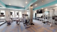 This fitness center offers a comprehensive workout environment designed to accommodate a wide range of exercise needs. The space is defined by a striking color scheme, with bold teal accent ceilings contrasting against crisp white and light gray walls, creating an energizing and modern atmosphere. Durable, patterned carpet tiles in neutral tones with blue geometric accents cover the floors throughout, providing both functionality and visual interest.

The layout is thoughtfully organized into distinct zones. The cardio area features multiple treadmills and elliptical machines, each equipped with individual monitors, positioned along a windowed wall that allows natural light to filter in. A strength training zone houses a variety of cable and resistance machines, including a multi-function cable station and a leg press machine, providing options for full-body workouts. A dedicated free weights area includes a complete dumbbell rack, with large wall mirrors running the length of that section to assist with form and technique.

A large flat-screen television is wall-mounted in a central location, visible from multiple areas of the room. A ceiling fan ensures air circulation throughout the space. Recessed lighting provides consistent illumination across the entire facility. A glass-paneled entrance door allows natural light into the entry area, which also features a fitness center policies placard. The space offers an open, well-organized feel that supports both individual and simultaneous multi-user workouts.