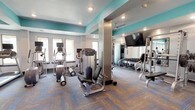 This fitness center offers a comprehensive workout environment designed to accommodate a wide range of exercise needs. The space is defined by a striking color scheme, with bold teal accent ceilings contrasting against crisp white and light gray walls, creating an energizing and modern atmosphere. Durable, patterned carpet tiles in neutral tones with blue geometric accents cover the floors throughout, providing both functionality and visual interest.

The layout is thoughtfully organized into distinct zones. The cardio area features multiple treadmills and elliptical machines, each equipped with individual monitors, positioned along a windowed wall that allows natural light to filter in. A strength training zone houses a variety of cable and resistance machines, including a multi-function cable station and a leg press machine, providing options for full-body workouts. A dedicated free weights area includes a complete dumbbell rack, with large wall mirrors running the length of that section to assist with form and technique.

A large flat-screen television is wall-mounted in a central location, visible from multiple areas of the room. A ceiling fan ensures air circulation throughout the space. Recessed lighting provides consistent illumination across the entire facility. A glass-paneled entrance door allows natural light into the entry area, which also features a fitness center policies placard. The space offers an open, well-organized feel that supports both individual and simultaneous multi-user workouts.