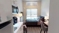 Located at 6105 Roosevelt Way NE in Seattle's vibrant Roosevelt neighborhood, this studio unit on the fourth floor offers a thoughtfully designed living space that maximizes every square foot. The open layout seamlessly integrates the sleeping, living, and kitchenette areas into a cohesive and functional environment. Large windows fitted with horizontal blinds allow abundant natural light to fill the room, creating a bright and welcoming atmosphere. The sleeping area features a full-sized bed with neutral-toned bedding, flanked by bedside lighting for a cozy ambiance. Hardwood-style flooring runs throughout, adding warmth and a clean, modern aesthetic. The compact kitchenette is outfitted with white cabinetry, a countertop sink, and space for small appliances, making everyday living convenient. A wall-mounted flat-screen TV and a dedicated desk area with seating provide both entertainment and workspace options within the unit. The neutral color palette of whites and grays throughout contributes to a calm, contemporary feel. This efficiently designed studio is well-suited for those seeking a modern urban living experience in one of Seattle's well-connected neighborhoods.