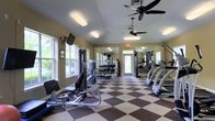 This single-floor fitness center is designed to support a comprehensive workout routine in a bright, welcoming environment. The space features a classic two-tone checkered floor that adds a timeless aesthetic while providing durable, easy-to-maintain surfacing throughout the exercise area. Large windows along the walls flood the room with abundant natural light and offer views of the surrounding greenery, creating an energizing atmosphere. Ceiling fans are mounted overhead to maintain comfortable airflow during workouts. The facility is well-stocked with a variety of strength-training equipment, including cable machines, a multi-station weight rack, and a full dumbbell rack, catering to users of all fitness levels. Cardio equipment such as treadmills is positioned to take advantage of the natural light and open sightlines. A large wall mirror enhances the sense of space and allows for proper form checks during exercise. The room is finished with warm, neutral-toned walls that contribute to the inviting ambiance. An exit door at the far end provides convenient access, and the overall layout ensures ample space to move freely between equipment, making this fitness center both functional and comfortable for daily use.