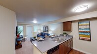 This single-floor residence at 400–480 43rd Ave SE in Puyallup, Washington offers a thoughtfully designed layout that balances comfort and functionality. The open-concept living and kitchen area serves as the heart of the home, where a peninsula with a double stainless steel sink and gray countertops connects seamlessly to the carpeted living room. Warm-toned wooden cabinetry pairs with stainless steel appliances — including a refrigerator, electric range, and built-in microwave — while hardwood-style flooring anchors the kitchen and entry. The living room features neutral walls, recessed beams, and access to an exterior door, allowing natural light to filter in alongside views of surrounding greenery. A flexible secondary room with a window and built-in shelving area offers space adaptable for an office or den. The bedroom is generously sized with plush carpet, a bright window overlooking outdoor foliage, and direct access to a spacious walk-in closet with built-in shelving. The bathroom is equipped with a full soaking tub and shower combination set on hardwood-style flooring. Throughout, the warm beige palette, consistent finishes, and practical layout create a cohesive and livable atmosphere.
