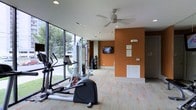 This fitness center offers a well-organized and functional workout environment spread across a single level. The space is anchored by floor-to-ceiling windows along one wall, flooding the interior with natural light and providing open views of the surrounding greenery outside — creating an energizing atmosphere for exercise. The patterned carpet flooring, featuring a subtle orange motif on a neutral background, adds a dynamic visual element underfoot while providing cushioning throughout the space. A bold accent wall painted in a warm terracotta orange serves as a striking focal point, complementing the overall color palette and giving the room a vibrant, motivating feel. The ceiling is outfitted with recessed lighting and a ceiling fan to maintain comfortable airflow during workouts. Equipment on display includes elliptical trainers, a stationary bike, and a cable strength training machine, catering to a range of fitness needs. A wall-mounted flat-screen television is positioned for easy viewing during cardio sessions. Mirrored panels along one section of the wall help to visually expand the space and allow for form checks during exercise. The layout is open and spacious, allowing for easy movement between equipment stations.