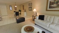 This single-floor residence offers a thoughtfully designed open-concept layout that blends the living and dining areas into one cohesive, inviting space. The living area is anchored by a plush white sofa accented with a dark decorative pillow, paired with a round white coffee table and a classic table lamp that casts a warm, welcoming glow. A dark leather ottoman adds a bold contrast against the neutral carpeted flooring, while framed artwork adorns the walls, lending a curated, personalized touch to the space.

Transitioning seamlessly from the living area, the dining space features a sleek glass-top table surrounded by dark chairs, complemented by a white sideboard beneath a pendant light. The combination of light tile flooring and warm beige walls throughout creates a bright and airy ambiance that feels both elegant and comfortable.

The neutral color palette, accented by warm lighting and carefully selected decor, gives the space a refined yet approachable character. The open flow between rooms enhances the sense of spaciousness, making this a versatile and functional home well-suited for both everyday living and entertaining.