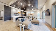 This thoughtfully designed single-floor amenity space offers a generous and well-appointed layout across three distinct zones. The main lounge area is anchored by large sectional seating arranged on a soft area rug, with light wood-look flooring extending throughout. Elevated ceilings with a recessed dark wood-planked tray detail and ceiling fans add warmth and architectural interest, while large sliding glass doors and full-height windows flood the space with natural light and provide direct access to an outdoor terrace.

A sleek wet bar runs along one wall, featuring dark wood-paneled cabinetry, stainless steel under-counter refrigeration, a white quartz countertop, and a wall-mounted fireplace display screen. A long white communal island nearby serves as a central gathering point, suspended beneath a sculptural pendant light. Adjacent to the lounge, a casual dining area includes round bistro-height tables and upholstered chairs, accented by organic wood wall sculptures that add a natural, artistic element.

A separate game and media room offers a ping pong table, multiple mounted televisions, and varied seating arrangements. Throughout the space, the cohesive palette of neutral tones, mixed textures, and curated décor creates a modern yet welcoming communal atmosphere.