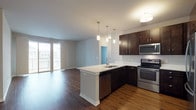 This single-floor residence is designed with a clean, contemporary aesthetic that prioritizes both functionality and style. The open-concept layout seamlessly connects the kitchen and living area, creating a spacious, cohesive environment well-suited for everyday living and entertaining.

The kitchen is a standout feature, outfitted with rich dark-espresso cabinetry that extends to the upper walls for ample storage. A full suite of stainless steel appliances includes a French-door refrigerator, electric range, over-the-range microwave, and dishwasher. The peninsula island features a white countertop and an undermount sink, with sleek pendant lighting suspended above. Warm hardwood-style flooring runs throughout the kitchen and living areas, complementing the modern finishes.

The living area is generously proportioned, featuring sliding glass doors that open to a private balcony, allowing natural light to fill the space. The bedroom offers plush neutral carpeting, a large window with horizontal blinds, and a calm, restful atmosphere.

The bathroom is well-appointed with dark cabinetry, a white countertop vanity, a large mirror with contemporary bar lighting, and matching hardwood-style flooring, maintaining the cohesive design language carried throughout the entire unit.