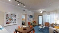 This single-floor residence showcases a bright and open layout that seamlessly connects the dining and living areas, creating a welcoming flow throughout the space. The dining area features a dark wood table complemented by bold orange metal chairs, adding a lively energy to the room. Track lighting on the ceiling provides focused illumination, while crown molding lends a polished, refined touch to the interior. Transitioning into the living area, a ceiling fan with a light fixture keeps the space comfortable, and sheer white curtains frame a window that allows natural light to filter in softly. A sleek, modern coffee table sits atop a rich blue area rug, anchoring the living space with color and texture. Wall-mounted décor and framed artwork are thoughtfully arranged throughout, contributing to the cheerful and curated aesthetic. A glimpse into the bedroom reveals a colorfully dressed bed and natural light from a window, maintaining the vibrant character found throughout the home. The textured ceilings and neutral wall tones provide a balanced backdrop that allows the bold furnishings and accents to take center stage, resulting in a cohesive and energetic living environment.