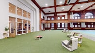 Located in the heart of Duluth, Minnesota, this remarkable single-floor space is a masterclass in the preservation of historic architecture. The centerpiece is a grand, soaring main hall featuring an indoor pickleball court, framed by rich, dark mahogany woodwork and ornate detailing that speaks to the building's storied past. A sweeping wraparound mezzanine balcony with intricate carved railings overlooks the main floor, supported by decorative arched windows that flood the space with natural light. The coffered ceiling, accented by dark wood beams and recessed lighting, adds dramatic vertical dimension to the hall. Surrounding the court, green turf areas offer flexible lounge and recreation zones, complete with modern seating arrangements and lawn games. Adjacent carpeted areas provide quieter gathering spaces with comfortable furnishings. A secondary area near the entrance features polished terrazzo flooring, pendant lighting, and communal tables, creating an inviting reception or common area. The hallway connects to accessible facilities, including an elevator, finished in clean, contemporary light wood paneling. Throughout, large, dark-framed windows offer views of the surrounding Duluth streetscape, seamlessly connecting this architecturally distinguished interior to its vibrant urban setting.