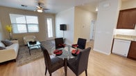 This single-floor residence offers a welcoming and functional open-concept layout that seamlessly connects the dining and living areas. The dining space is anchored by a round glass-top table surrounded by four dark upholstered chairs, creating an elegant yet casual setting for everyday meals and gatherings. A wall-mounted flat-screen TV serves as a central focal point between the two zones, adding a modern, streamlined touch to the design.

The living area features a comfortable seating arrangement set atop a textured area rug, complemented by a glass coffee table. Natural light flows generously into the space through multiple windows, enhancing the airy and open atmosphere. A ceiling fan with an integrated light fixture keeps the space comfortable year-round while adding a classic design element overhead.

Warm hardwood-style flooring runs throughout, tying the entire space together with a cohesive and clean aesthetic. Crisp white walls and neutral tones create a bright, inviting ambiance that feels both spacious and easy to personalize. A hallway visible from the main living area leads to additional rooms, suggesting a practical and well-organized floor plan. The front entryway, framed by a windowed door, adds an additional source of natural light and a welcoming sense of arrival.