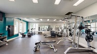 Located at 2018 Cunningham Dr in Hampton, Virginia, this fitness center offers a well-equipped, single-floor workout facility designed to support a full range of exercise routines. The space features a clean, modern aesthetic with white walls accented by bold teal feature walls that add energy and visual interest to the environment. Large windows on multiple sides flood the room with natural light and provide views of the surrounding greenery, creating an inviting and motivating atmosphere. Durable rubber flooring covers the workout area, ensuring safety and comfort during exercise. A full-length wall mirror expands the sense of space and allows for form checking during workouts. The cardio section is well-stocked with treadmills, ellipticals, and stationary bikes, while the strength training area includes a multi-function cable machine, a squat/power rack with weight plates, a flat adjustable workout bench, and a dumbbell rack with a wall-mounted flat-screen TV. Overhead fluorescent lighting ensures the space remains bright at all hours. Multiple entry and exit points, including glass-panel French doors connecting to an adjacent corridor and an exterior door leading outdoors, provide convenient access throughout. The overall layout is open and thoughtfully organized, offering ample room to move freely between stations.