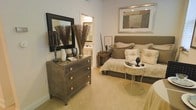 This single-floor space showcases a thoughtfully designed bedroom that exudes understated elegance through a carefully curated neutral color palette. The room is anchored by a generously sized upholstered bed with a tufted headboard in warm taupe, dressed in layered cream and champagne bedding that adds depth and texture to the space. Soft, plush wall-to-wall carpeting in a warm beige tone runs throughout, contributing to the room's cozy and refined ambiance.

A distinctive metallic three-drawer dresser serves as a functional focal point along one wall, topped with decorative accents including white ceramic vases, a black-framed mirror, and sculptural dried botanical arrangements that add organic warmth. A framed abstract artwork in muted tones is positioned above the bed, complementing the room's sophisticated aesthetic.

A delicate wrought-iron side table sits beside the bed, offering a charming contrast to the heavier furniture pieces. An en-suite bathroom is accessible directly from the bedroom, featuring warm lighting and clean finishes. The overall ambiance of the space is serene, cohesive, and designed for comfort and relaxation.