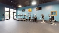 This fitness center occupies a single open floor and is designed to accommodate a comprehensive range of workout activities. The space is immediately distinguished by its bold teal walls, which are complemented by a dark, exposed ceiling that adds an industrial character while maintaining a spacious, airy feel. Durable speckled rubber flooring runs throughout, providing a practical and safe surface suited to high-traffic exercise use.

The cardio zone features a row of treadmills, elliptical machines, and upright stationary bikes, all positioned along one wall beneath wall-mounted flat-screen televisions, allowing users to stay entertained during their workouts. Natural light filters in through a glass entry door, brightening this section of the space.

The strength training area includes a multi-station cable machine, a variety of selectorized resistance machines, and a dumbbell rack stocked with free weights. Gold-framed mirrors are strategically placed throughout the facility, enhancing the sense of depth and allowing users to monitor their form. A dedicated open-floor zone offers additional space for stretching or functional training. Structural columns are integrated into the layout, adding to the industrial aesthetic of the overall space.