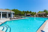 Located at 915 Lovvorn Rd in Carrollton, Georgia, this outdoor amenity area offers a well-appointed resort-style experience centered around a generously sized swimming pool with clear turquoise water. The pool is framed by a broad concrete deck outfitted with multiple lounge chairs, providing ample space for sunbathing and relaxation. A built-in basketball hoop within the pool adds a recreational element for active enjoyment.

Surrounding the pool, shaded pergola-style cabanas with curtains offer a more private retreat, while umbrella-covered seating areas provide additional options for outdoor dining and lounging. A built-in grill station with a stone surround makes the space ideal for outdoor gatherings. Classic lamp post lighting is positioned throughout the deck, enhancing both the aesthetic and functionality of the area into the evening hours.

Adjacent to the pool area stands a two-story clubhouse building with navy blue board-and-batten siding and large upper-level windows, lending a charming, cottage-inspired architectural character. A separate utility shed in matching navy blue siding complements the overall design. The entire area is enclosed by a wrought iron fence, set against a backdrop of mature trees that provide natural privacy and greenery, creating a welcoming and well-maintained community amenity space.