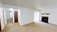 This single-floor residence offers a well-planned layout within a 1-bedroom, 1-bathroom configuration. The living area serves as the central gathering space, featuring wall-to-wall carpet flooring that adds warmth underfoot. A standout feature of the room is the classic fireplace, framed by a sleek dark surround and topped with a simple mantel shelf, providing both a functional heat source and a focal point for the space. Large vertical blinds cover a full-length window, allowing for adjustable natural light throughout the day while maintaining privacy. The entryway is defined by a solid wood-toned front door, providing a clean transition from the exterior into the living space. The white walls throughout lend a bright, neutral backdrop that makes the space feel open and easy to personalize. The single-floor layout ensures convenient, step-free living with all areas readily accessible. Overall, this residence combines practical design with comfortable finishes, making it a functional and inviting home.
