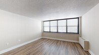 This single-floor unit offers a thoughtfully arranged layout that maximizes its available space. The main living area is a standout feature, characterized by a wide bank of floor-to-ceiling windows fitted with horizontal blinds, allowing generous natural light to fill the room. Warm wood-look laminate flooring runs throughout the space, complemented by neutral beige walls and crisp white baseboards. A textured popcorn ceiling adds a classic finishing detail overhead. A wall-mounted baseboard heating unit ensures climate comfort within the living area.

The compact kitchen is efficiently designed, featuring white cabinetry with brushed hardware, granite-look countertops, a double-basin sink, and a white gas range with oven. A full-size refrigerator and under-cabinet space round out the kitchen's functionality. The entry door opens directly adjacent to the kitchen, creating a convenient and direct access point into the unit.

The bathroom presents light blue square wall tiles surrounding a full soaking tub with a showerhead and grab bar. Mosaic tile flooring in coordinating tones adds visual interest, while wire shelving along one wall provides practical storage. The overall ambiance of the unit is clean, functional, and filled with natural light.