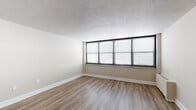 This single-floor unit offers a thoughtfully arranged layout that maximizes its available space. The main living area is a standout feature, characterized by a wide bank of floor-to-ceiling windows fitted with horizontal blinds, allowing generous natural light to fill the room. Warm wood-look laminate flooring runs throughout the space, complemented by neutral beige walls and crisp white baseboards. A textured popcorn ceiling adds a classic finishing detail overhead. A wall-mounted baseboard heating unit ensures climate comfort within the living area.

The compact kitchen is efficiently designed, featuring white cabinetry with brushed hardware, granite-look countertops, a double-basin sink, and a white gas range with oven. A full-size refrigerator and under-cabinet space round out the kitchen's functionality. The entry door opens directly adjacent to the kitchen, creating a convenient and direct access point into the unit.

The bathroom presents light blue square wall tiles surrounding a full soaking tub with a showerhead and grab bar. Mosaic tile flooring in coordinating tones adds visual interest, while wire shelving along one wall provides practical storage. The overall ambiance of the unit is clean, functional, and filled with natural light.