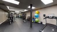 Located at 919 N Stone Ave in Tucson, Arizona, this fitness center offers a comprehensive, single-floor workout facility designed to accommodate a wide range of exercise needs. The space features durable dark rubber flooring throughout, providing a safe and functional surface for all types of training activities.

The cardio area is equipped with multiple treadmills, elliptical trainers, and stationary and spin bikes, all arranged along one side of the room for easy access. A dedicated strength training zone includes a multi-station cable machine, an adjustable weight bench, and a well-stocked dumbbell rack with a full range of free weights. Medicine balls and stability balls of varying sizes are neatly organized on wall-mounted and freestanding racks, supporting functional fitness and flexibility training.

The facility is illuminated by large recessed panel lighting in a drop-tile ceiling, ensuring bright and even light across the entire floor. A floor-to-ceiling glass partition wall with black framing separates the gym from an adjacent common area, allowing natural light to filter in and creating an open, connected feel. Full-length mirrors in the strength training section enhance the sense of space and allow users to monitor their form. The overall design is clean, modern, and purposefully organized to support an efficient and effective workout experience.