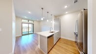 This thoughtfully designed one-bedroom, three-bathroom residence offers a efficient and modern living experience across a single floor. The open-concept living and kitchen area is anchored by a striking island topped with elegant veined quartz countertops, complemented by a stainless steel sink and dishwasher. The kitchen is fully equipped with gray shaker-style cabinetry, a subway tile backsplash, and a full suite of stainless steel appliances including a refrigerator, range, and built-in microwave. Warm hardwood-style flooring flows throughout the main living area, which is bathed in natural light from large sliding glass doors that open to a private balcony. Pendant lighting above the island adds a refined touch to the space. The bedroom features plush carpet flooring, a ceiling fan, a bright window with outdoor views, and access to a spacious walk-in closet with built-in shelving and hanging rods. The bathroom is finished with large-format tile flooring, gray vanity cabinetry, white countertops, and a soaking tub with tiled surround. Clean, neutral tones and modern finishes throughout give the space a cohesive and contemporary aesthetic.