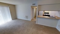 This single-floor residence features an open-concept layout that connects the main living area to the kitchen, creating a functional and cohesive living space. The living area is finished with wall-to-wall beige carpeting, offering a comfortable and neutral foundation throughout. White walls and a textured ceiling keep the space feeling clean and straightforward, while a ceiling-mounted HVAC vent and a floor-level return air vent indicate a central climate control system for year-round comfort.

A half-wall partition separates the living area from the kitchen, which is equipped with a refrigerator, a gas range, and cabinetry finished in a dark wood tone. The countertop features a light wood laminate surface, providing practical workspace. This pass-through design between the kitchen and living area promotes an open, connected feel.

A doorway visible in the background leads to a hallway that provides access to additional rooms, including what appears to be a closet with bi-fold doors. The overall layout is straightforward and efficiently designed, making practical use of the available space. The neutral palette throughout lends itself well to a variety of furnishing styles.