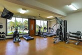 This single-story fitness center in Opelika, Alabama offers a well-equipped and functional workout environment designed to support a variety of exercise routines. The space features polished concrete flooring throughout, giving it a clean, durable, and modern aesthetic that is easy to maintain.

The main workout area is generously proportioned, accommodating a diverse selection of cardio and strength training equipment, including treadmills, elliptical machines, a multi-station cable weight machine, and a rack of free weights. Large windows along the walls allow abundant natural light to fill the space while providing views of the surrounding outdoor area, including what appears to be a pool and landscaped grounds. A wall-mounted television is positioned for visibility during workouts.

Exposed ductwork along the ceiling adds an industrial character to the space, complemented by flush and overhead lighting fixtures. A ceiling fan aids in air circulation throughout the facility.

The entrance area features a dark-framed glass door flanked by sidelight windows, offering a welcoming entryway with clear outdoor views. A built-in water fountain is conveniently located near the entry. Yoga mats stored near the entrance indicate the space also accommodates stretching and floor-based exercise.

A dedicated restroom, clearly marked with accessibility signage, is accessible from the central corridor area, ensuring full convenience for users of the facility.